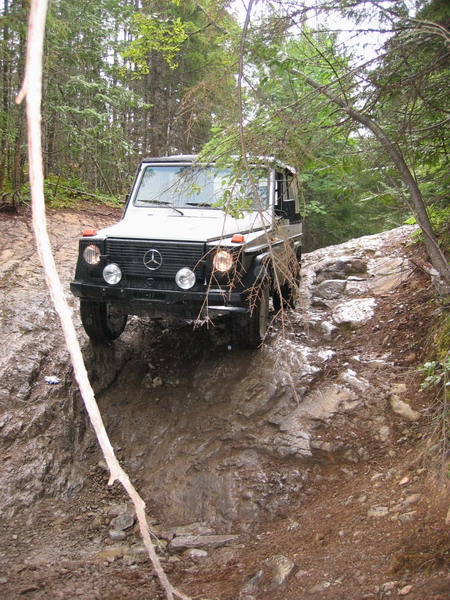 1985 Ford Bronco - A white Mercedes-Benz G-Wagen is navigating a steep, muddy, and rocky off-road trail through a forest.