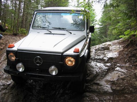 1985 Ford Bronco - A white Mercedes-Benz G-Wagen is navigating a rocky, wet, off-road trail in a forest. The vehicle's headlights and turn signals are illumin…