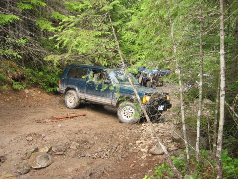 1985 Ford Bronco - A dark blue Jeep Cherokee XJ is parked on a muddy, wooded trail with other vehicles visible in the background. The SUV is angled on the une…