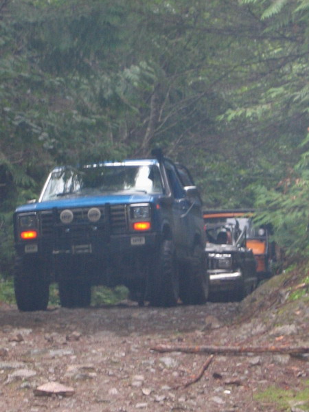 1985 Ford Bronco - A blue Ford Bronco is driving on a dirt road through a forest, pulling another off-road vehicle behind it. The Bronco has large tires and a…