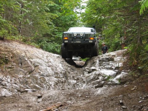 1985 Ford Bronco - A lifted, dark-colored Ford Bronco navigates a steep, rocky, and muddy off-road trail through a dense forest.