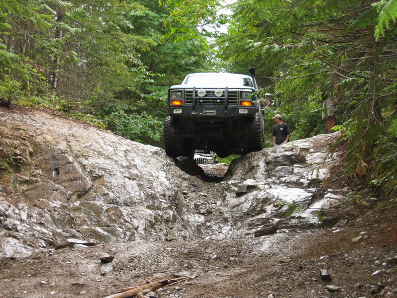 Lifted Ford Bronco driving over rocks on a muddy forest trail.