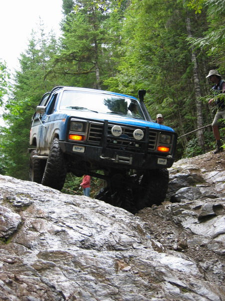 1985 Ford Bronco - A lifted blue Ford Bronco is navigating a rocky, off-road trail with trees on either side. The vehicle has large tires, a snorkel, and auxi…