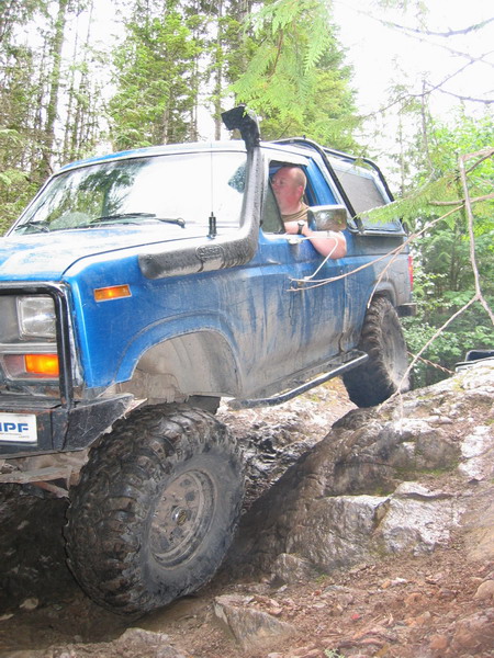 1985 Ford Bronco - A blue Ford Bronco is navigating a rocky, off-road trail with a driver visible behind the wheel. The vehicle has large, aggressive tires an…
