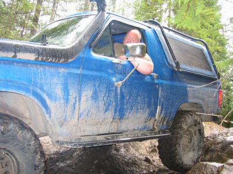 1985 Ford Bronco - A blue, mud-covered Ford Bronco is navigating a rocky trail with a snorkel visible on the driver's side. The driver is visible through the…