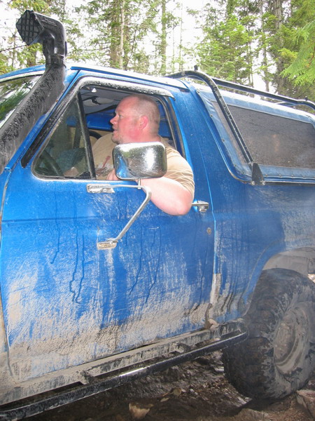 1985 Ford Bronco - A man with a shaved head looks out the driver's side window of a mud-splattered blue SUV. The vehicle has a snorkel attached to the A-pilla…