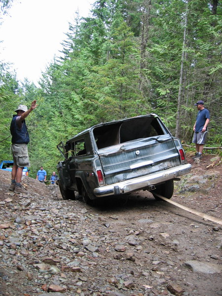 1985 Ford Bronco - A dark green SUV is being pulled up a rocky, wooded hill by a tow strap. Two men are visible, one directing the tow and another standing ne…