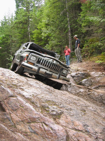 1985 Ford Bronco - A damaged Jeep Cherokee is being winched up a steep, rocky incline in a wooded area. Two people are standing on the trail watching the reco…
