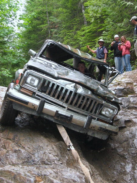1985 Ford Bronco - A heavily damaged Jeep Cherokee is tilted precariously on a rocky incline, with a tow strap attached to its front bumper. Several people wa…