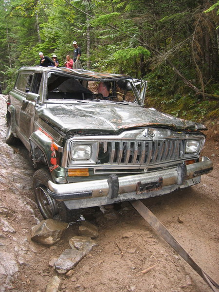 1985 Ford Bronco - A heavily damaged green Jeep Cherokee is stuck in mud on a wooded trail, with its roof caved in and doors open. Several people are standing…