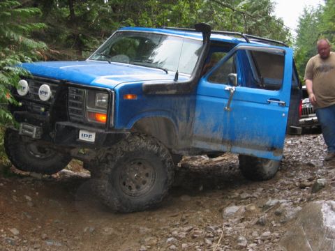 1985 Ford Bronco - A bright blue, lifted Ford Bronco with large off-road tires navigates a rocky, muddy trail. The driver's side door is open, revealing the i…