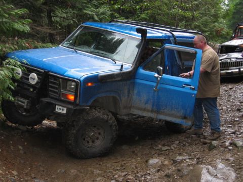 1985 Ford Bronco - A blue Ford Bronco is parked on a muddy, rocky trail with its driver's side door open. The vehicle has a snorkel, off-road tires, and auxil…