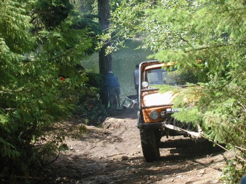 1985 Ford Bronco - An orange off-road vehicle is parked on a dirt path next to a lake, with trees obscuring the view. Two people are visible near the water's…
