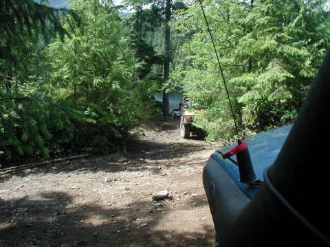 1985 Ford Bronco - A dirt path leads through a dense forest towards a body of water, with two off-road vehicles visible. The foreground shows the hood and sno…