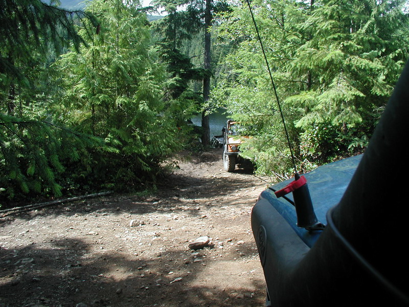 Two off-road vehicles on a dirt path in a forest near water. View from inside a dark vehicle.