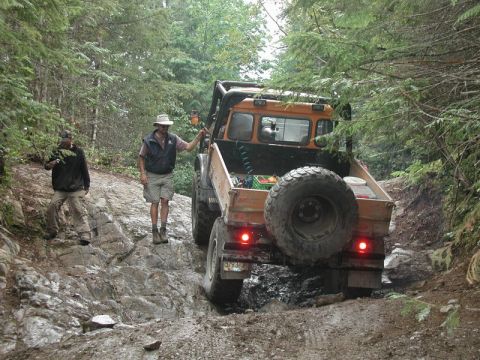 1985 Ford Bronco - An orange off-road truck with large tires navigates a rocky, muddy trail in a forest. Two men are standing near the truck, one appearing to…