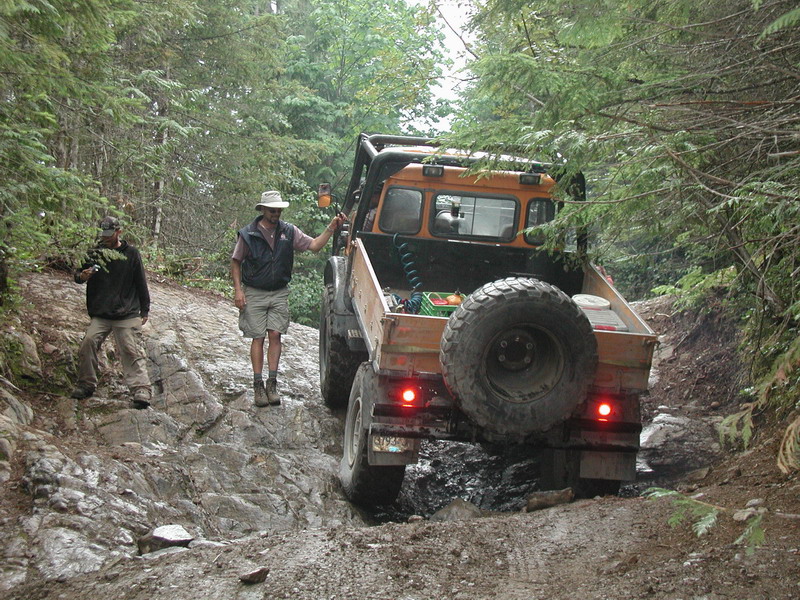 Orange off-road truck on a rocky, muddy forest trail with two men.