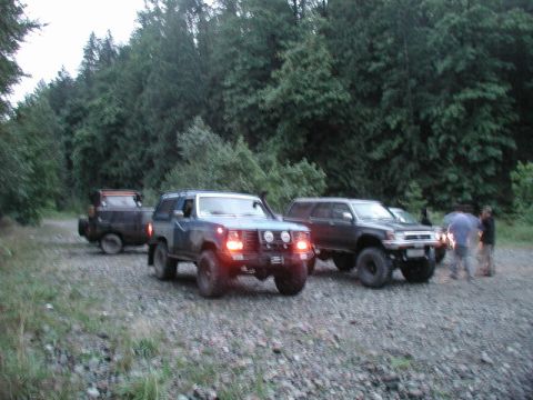 1985 Ford Bronco - Three off-road vehicles are parked on a gravel road in a forest. The closest vehicle is a dark blue SUV with large tires and auxiliary ligh…