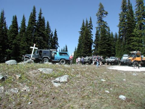 1985 Ford Bronco - A group of off-road vehicles are parked on a grassy, rocky hilltop with scattered yellow flowers and tall evergreen trees in the background…