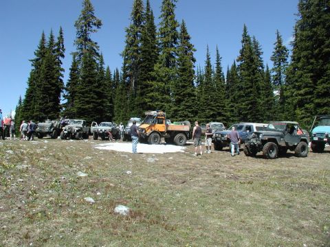 1985 Ford Bronco - A group of off-road vehicles are parked in a clearing surrounded by tall evergreen trees. A bright orange six-wheeled truck is prominent in…
