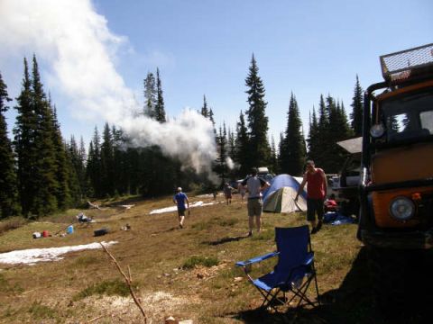 1985 Ford Bronco - A group of people are camping in a wooded area with a large orange off-road vehicle parked on the right side of the frame. Smoke is rising…