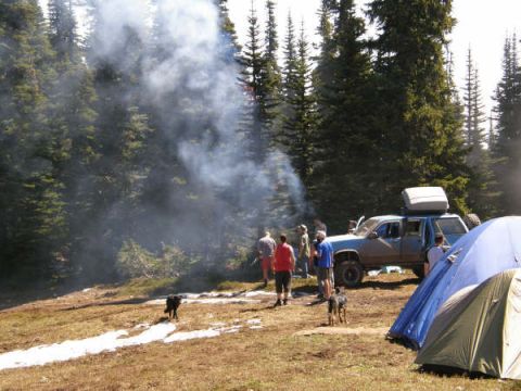 1985 Ford Bronco - A blue SUV is parked in a wooded area next to a campsite with tents and people. Smoke is rising from a fire in the background.
