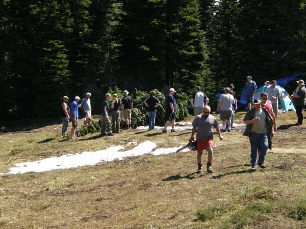 A group of people in a wooded area with tents, one man holds a chainsaw.