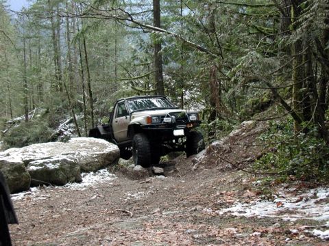 1985 Ford Bronco - A lifted, white pickup truck with a flatbed is navigating a rocky, wooded trail with patches of snow on the ground. The truck is angled tow…