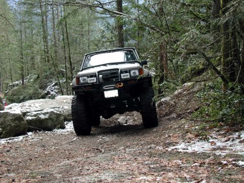 1985 Ford Bronco - A lifted, modified off-road SUV navigates a snowy, wooded trail with large tires and a winch on the front bumper.