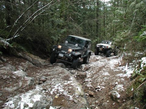 1985 Ford Bronco - Two off-road vehicles navigate a rocky, snow-dusted forest trail. The lead vehicle is a dark-colored Jeep Wrangler, while the second vehicl…