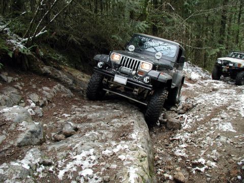 1985 Ford Bronco - A dark colored Jeep Wrangler is navigating a rocky, off-road trail with patches of snow on the ground. Another off-road vehicle is visible…