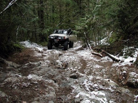 1985 Ford Bronco - A lifted, light-colored SUV navigates a rocky, wooded trail with patches of snow on the ground. The vehicle is positioned on the right side…