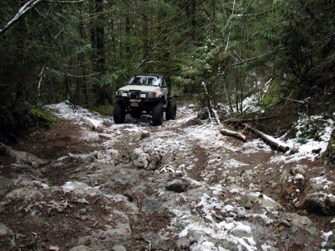 1985 Ford Bronco - A lifted, tan off-road SUV navigates a rocky, snow-dusted trail through a dense forest.