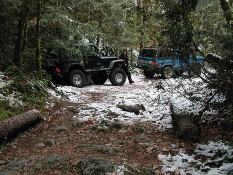 1985 Ford Bronco - Two off-road vehicles are parked on a snow-dusted forest trail. A black Jeep Wrangler is on the left, and a blue Ford Bronco is to its righ…