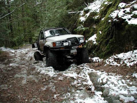 1985 Ford Bronco - A white pickup truck with large off-road tires navigates a rocky, snow-dusted forest trail. The truck features a winch and auxiliary lights…