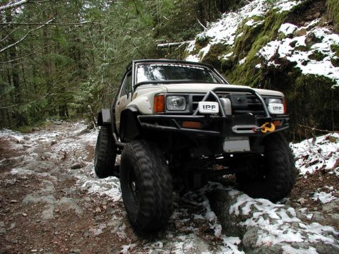 1985 Ford Bronco - A modified, lifted Toyota pickup truck with large off-road tires navigates a rocky, snow-dusted forest trail.