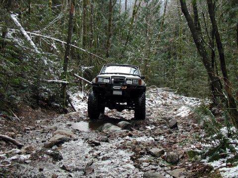 1985 Ford Bronco - A lifted, white 1980s SUV navigates a rocky, wooded trail with patches of snow. The vehicle is equipped with off-road tires and auxiliary l…