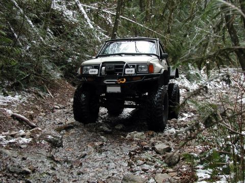 1985 Ford Bronco - A lifted, off-road modified pickup truck with large tires navigates a rocky, wooded trail with patches of snow on the ground.
