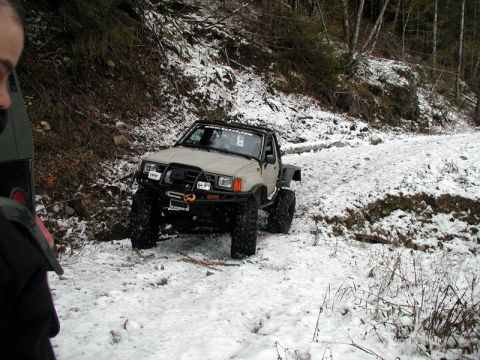 1985 Ford Bronco - A tan off-road pickup truck with large tires is navigating a snowy, wooded trail. The vehicle has a winch mounted on the front bumper and a…