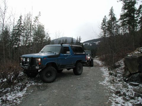1985 Ford Bronco - A blue Ford Bronco and a dark colored Jeep are parked on a gravel road with patches of snow. The Bronco is equipped with a snorkel and a br…