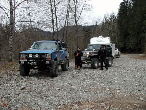 1985 Ford Bronco - A blue Ford Bronco and a black Jeep Wrangler with a trailer are parked on a gravel road in a wooded area. Two people stand near the Jeep.
