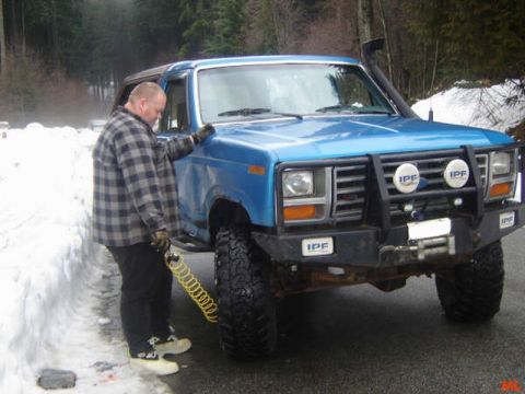 1985 Ford Bronco - A man in a plaid shirt stands next to a blue Ford Bronco in a snowy environment. He is holding a yellow coiled air hose near the front tire.