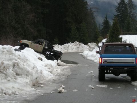 1985 Ford Bronco - A blue Ford Bronco is parked on a wet, paved road next to a large pile of snow. In the background, a tan pickup truck is partially stuck in…