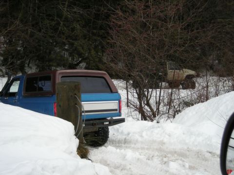 1985 Ford Bronco - A blue Ford Bronco with a brown top is parked in the snow next to a wooden post. Another off-road vehicle is visible in the background.