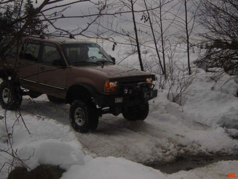1985 Ford Bronco - A lifted brown SUV with large tires drives through a snowy, wooded area, its headlights on.
