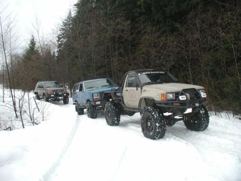 1985 Ford Bronco - Three modified off-road vehicles drive in single file on a snow-covered trail through a forest. The lead vehicle is a tan pickup truck with…