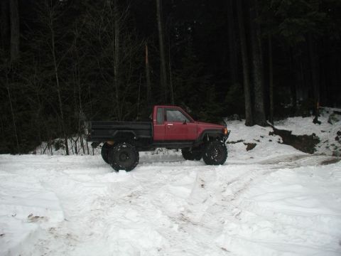 1985 Ford Bronco - A red flatbed pickup truck with large off-road tires is parked in a snowy forest clearing. The truck has a black flatbed and appears to be…