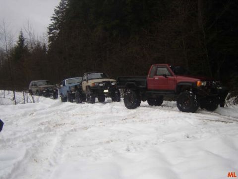 1985 Ford Bronco - Four modified off-road vehicles are parked on a snow-covered path in a wooded area. A red pickup truck with large tires is in the foregroun…
