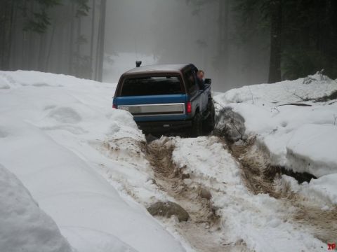 1985 Ford Bronco - A blue Ford Bronco with a brown roof drives through a snowy, wooded area, leaving tracks in the mud and snow. The driver can be seen lookin…