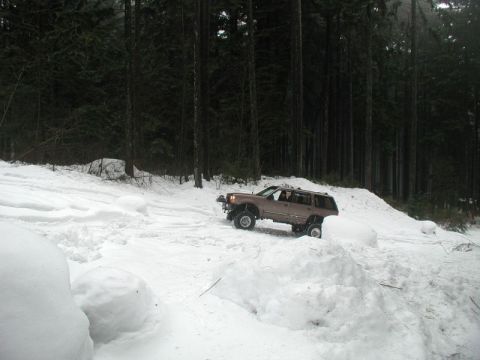 1985 Ford Bronco - A brown Ford SUV navigates a snow-covered trail with large snow mounds and a dense forest in the background.
